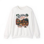 White Cozy Cabin Reading Retreat sweatshirt with a glowing log cabin in the pines, stacks of books and a crackling fire, front view
